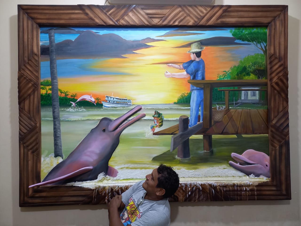 Cena de um pôr do sol vibrante em tons de amarelo e laranja sobre um rio. À direita, um homem sobre um trapiche de madeira pescando um tucunaré enquanto um boto-cor-de-rosa que emerge da água com a boca aberta. Outro boto salta ao fundo, próximo a um barco regional de dois andares. A moldura é feita de talas de miriti trançadas em padrões geométricos.